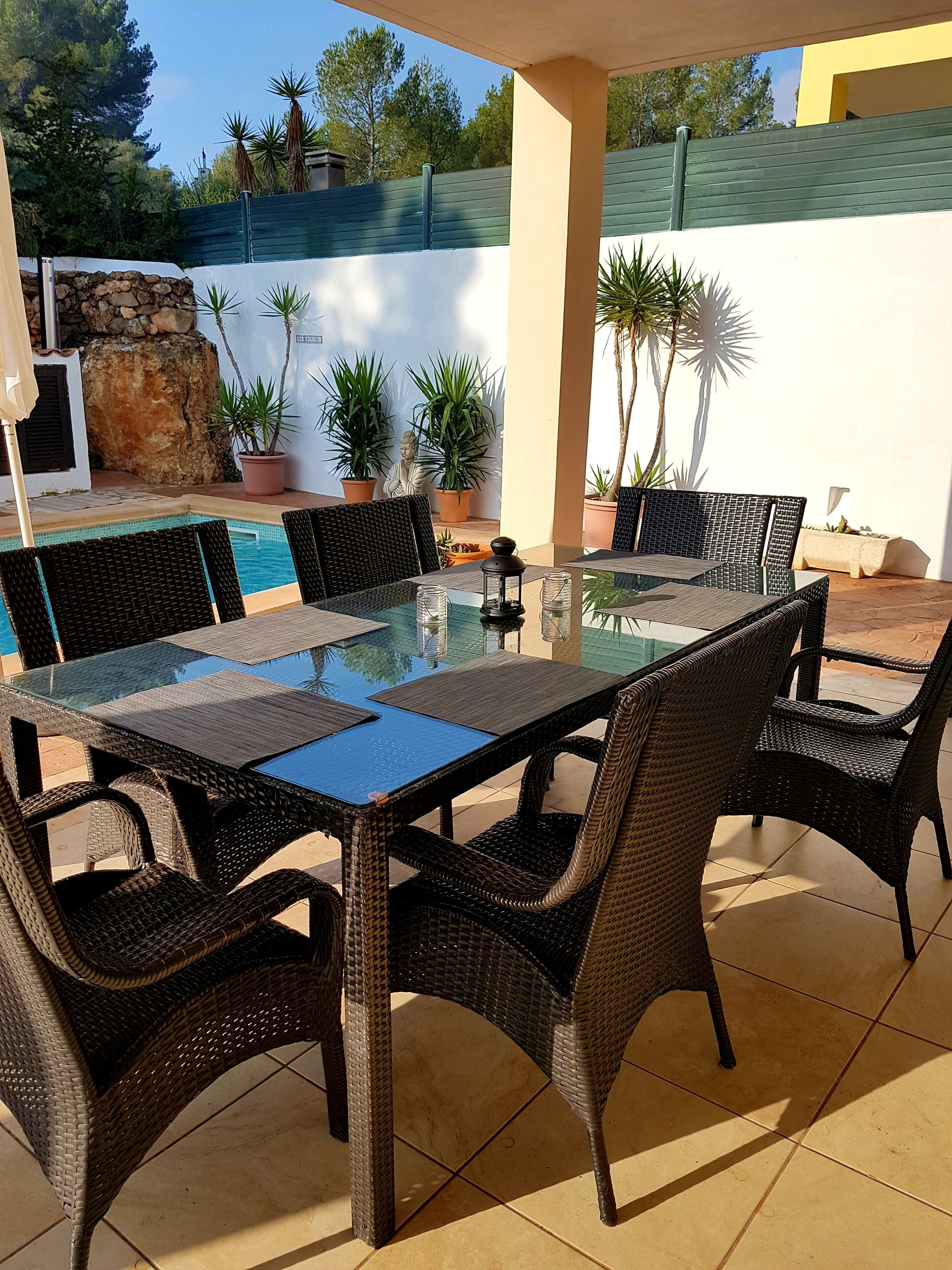 En udendørs terrasse med et bord med glasplade, omgivet af fletstole, en stenvæg og krukker med planter.