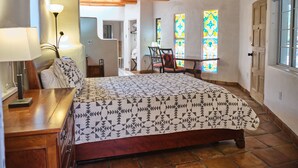 A bedroom with a wooden ceiling, a bed with a patterned bedspread, a nightstand, a lamp, and a chair. There are two windows with stained glass, a door, and a hallway leading to another room.