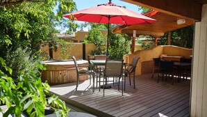 An outdoor patio with a wooden deck, a hot tub, and outdoor furniture under a red umbrella.