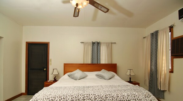 Quarto com cama, duas mesas de cabeceira, ventoinha no teto, porta e cortinas.