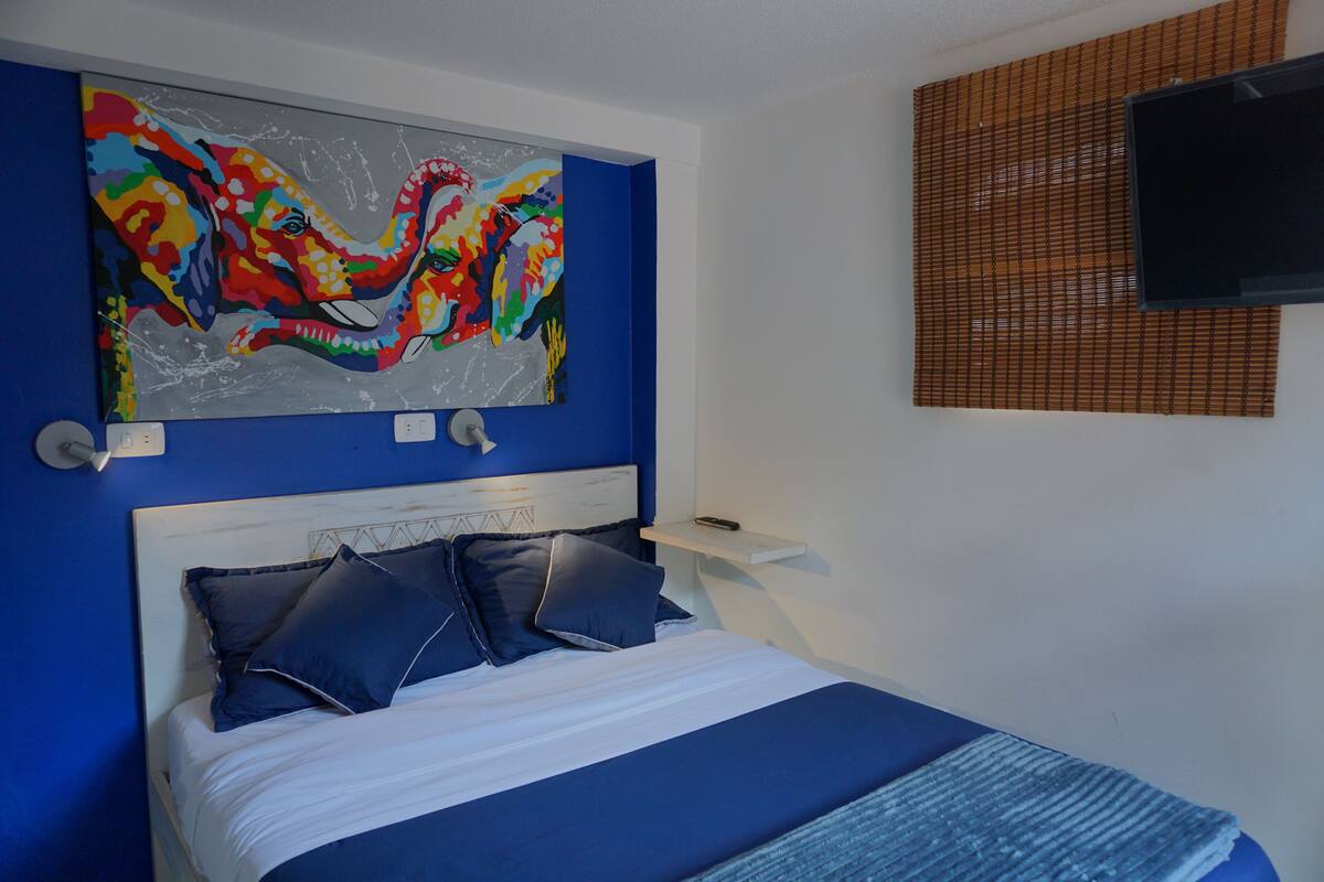 Une chambre à coucher avec un lit, un tableau abstrait aux couleurs vives, un téléviseur et une fenêtre avec des stores.