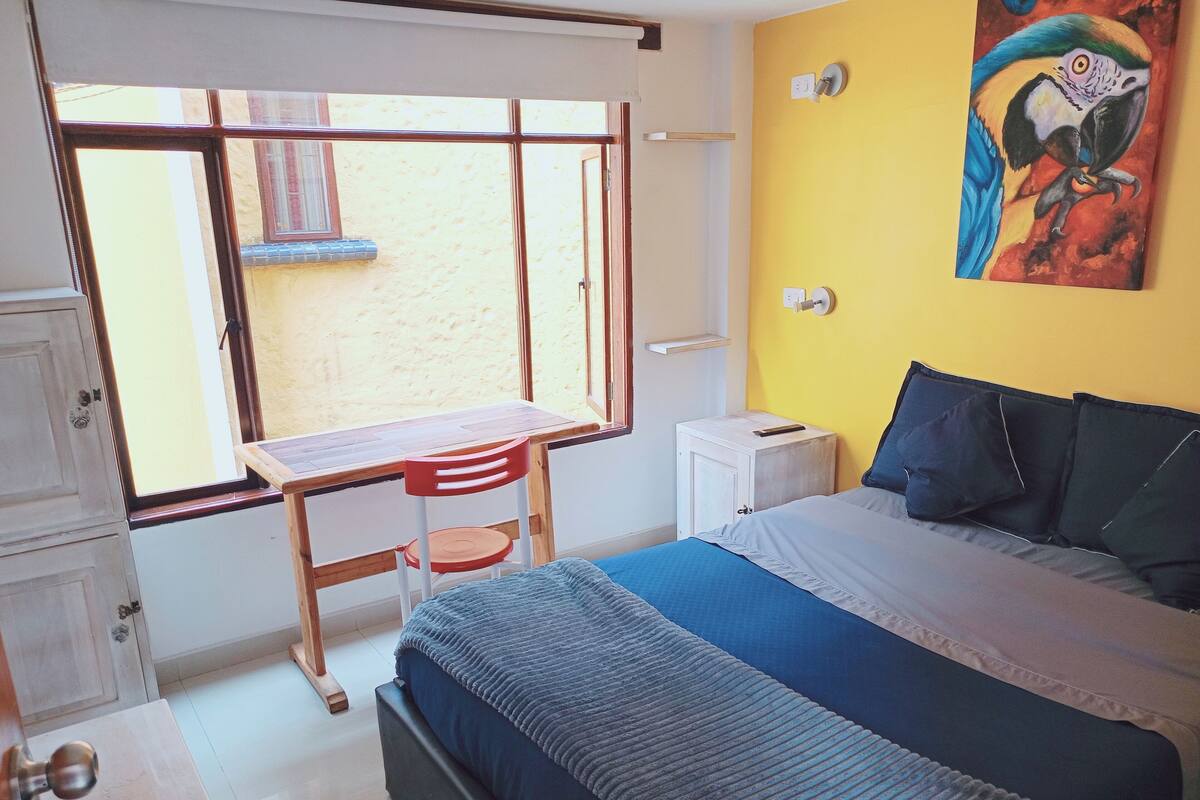 Une chambre à coucher avec un lit, un bureau et une chaise. Il y a une fenêtre et un tableau au mur.