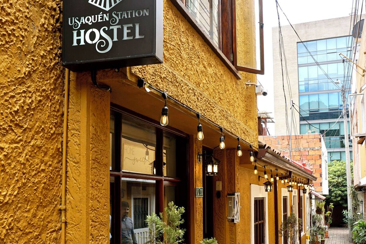 Une auberge de jeunesse avec une enseigne indiquant « USAQUEN Station Hostel », et une ruelle étroite agrémentée de plantes en pot et de quelques tables et chaises à l’extérieur.