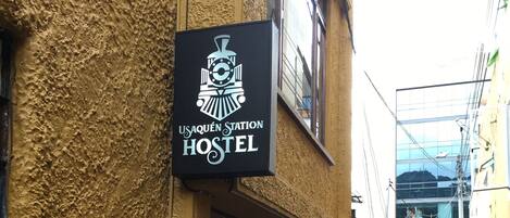 A hostel with a sign reading "USAQUEN Station Hostel" on a textured yellow wall.