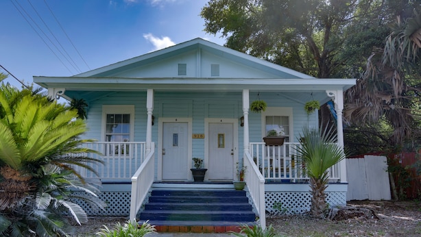 A light blue house with a porch, numbered doors, and a small garden with palm trees.