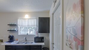 A compact kitchen with a sink, a microwave, and a coffee maker. There are two shelves above the sink and a trash can next to the door.