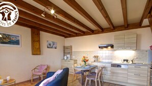 A cozy living room with a blue sofa, a dining table with chairs, and a kitchen area with cabinets and a stove. - Cottage Les Hortensias in Pralong - Charm and Comfort for 4 Guests with Internet (Pralong)
