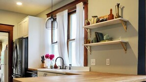 A kitchen with a wooden island, two bar stools, a refrigerator, and a hanging light fixture.