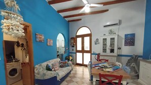 A room with a blue wall, a ceiling fan, a sofa, a dining table with chairs, and a kitchen area visible through an arched doorway. - Ludovica's dream house  (Bagheria)