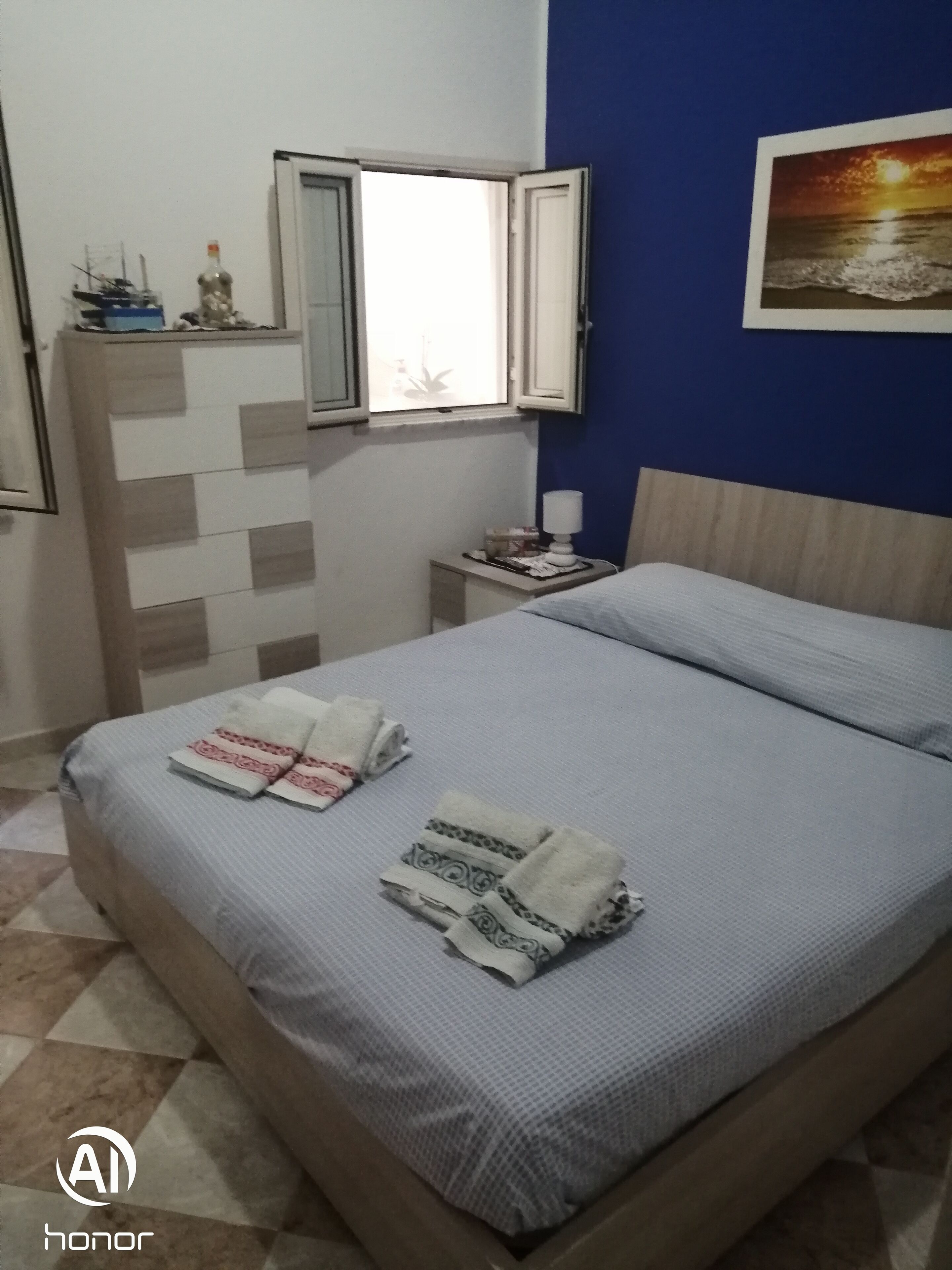 A neatly made bed with towels, a dresser with a mirror, a small table with a lamp, and a painting on the wall.