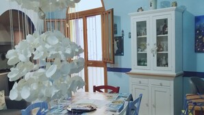 A dining area with a table set for four, surrounded by chairs. A white cabinet with glass doors is to the right, and a wooden-framed window is behind the table. - Ludovica's dream house  (Bagheria)