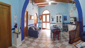 A living room with a checkered floor, a sofa, a dining table with chairs, and a kitchen area with cabinets and a hanging light fixture. - Ludovica's dream house  (Bagheria)