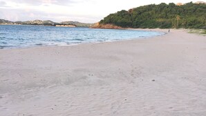 A wide sandy beach with a calm sea and a forested hill in the background.