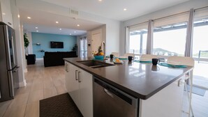A modern kitchen with a central island, a dishwasher, and a sink. There is a dining area with a table and chairs, and a large window providing a view.