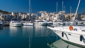 Un puerto deportivo con varias embarcaciones amarradas, rodeado de edificios blancos con balcones y palmeras.