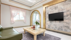 A modern living room with a green sofa, a wooden coffee table with a vase and bottles, a flat-screen TV mounted on a textured wall, and a large window with pink curtains. - Gyeongju Woorinuri Pension (Gyeongju)