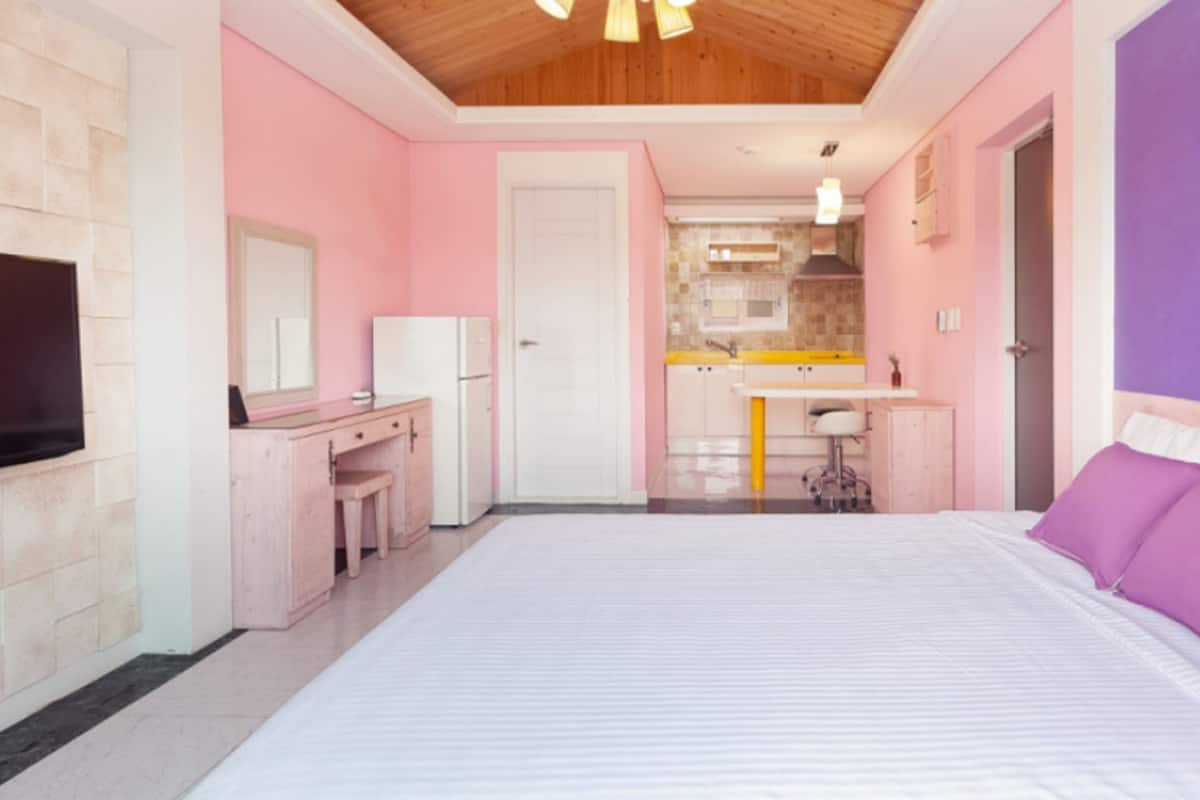 A bedroom with a large bed, a desk, and a kitchenette in the background.