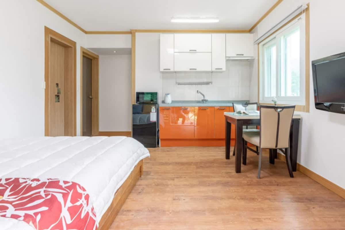 A compact hotel room with a bed, a desk with a chair, a television, and a kitchenette in the background.