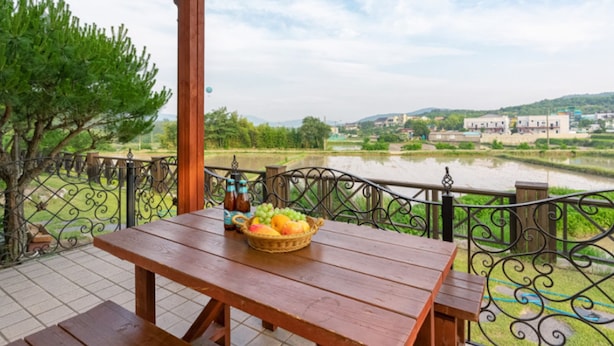 A wooden table with a basket of fruit and bottles on a balcony overlooking a river and a distant building. - Gyeongju Woorinuri Pension (Gyeongju)