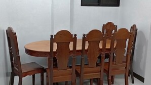 Uma área de refeições com uma mesa de madeira redonda e seis cadeiras, uma pequena janela e paredes brancas.
