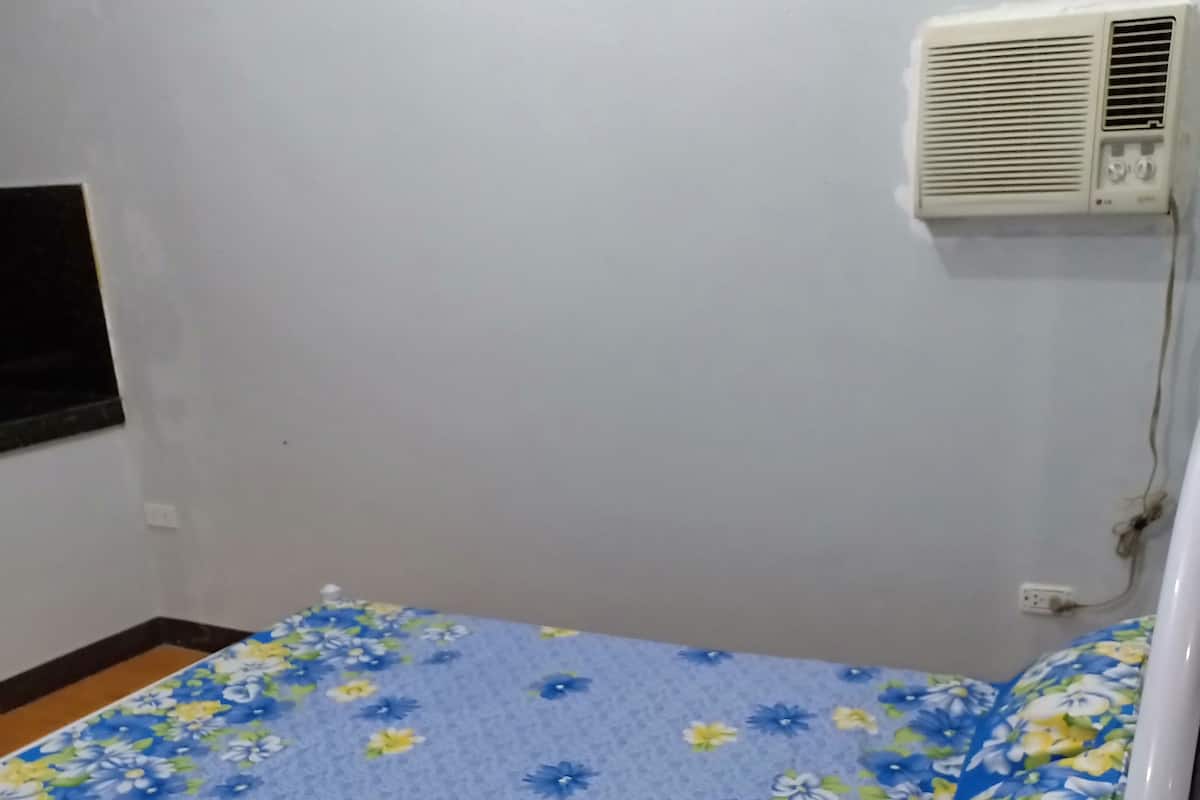 A small, single-bed room with a floral-patterned bedspread, a white wall, and a wall-mounted air conditioner.