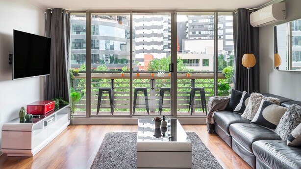 A modern living room with a sofa, a coffee table, and a TV. Large windows offer a view of the cityscape. - Southbank Boulevard Melbourne (Southbank)