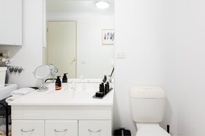 A bathroom with a white sink, mirror, and toiletries. There is a white toilet and a door in the background.