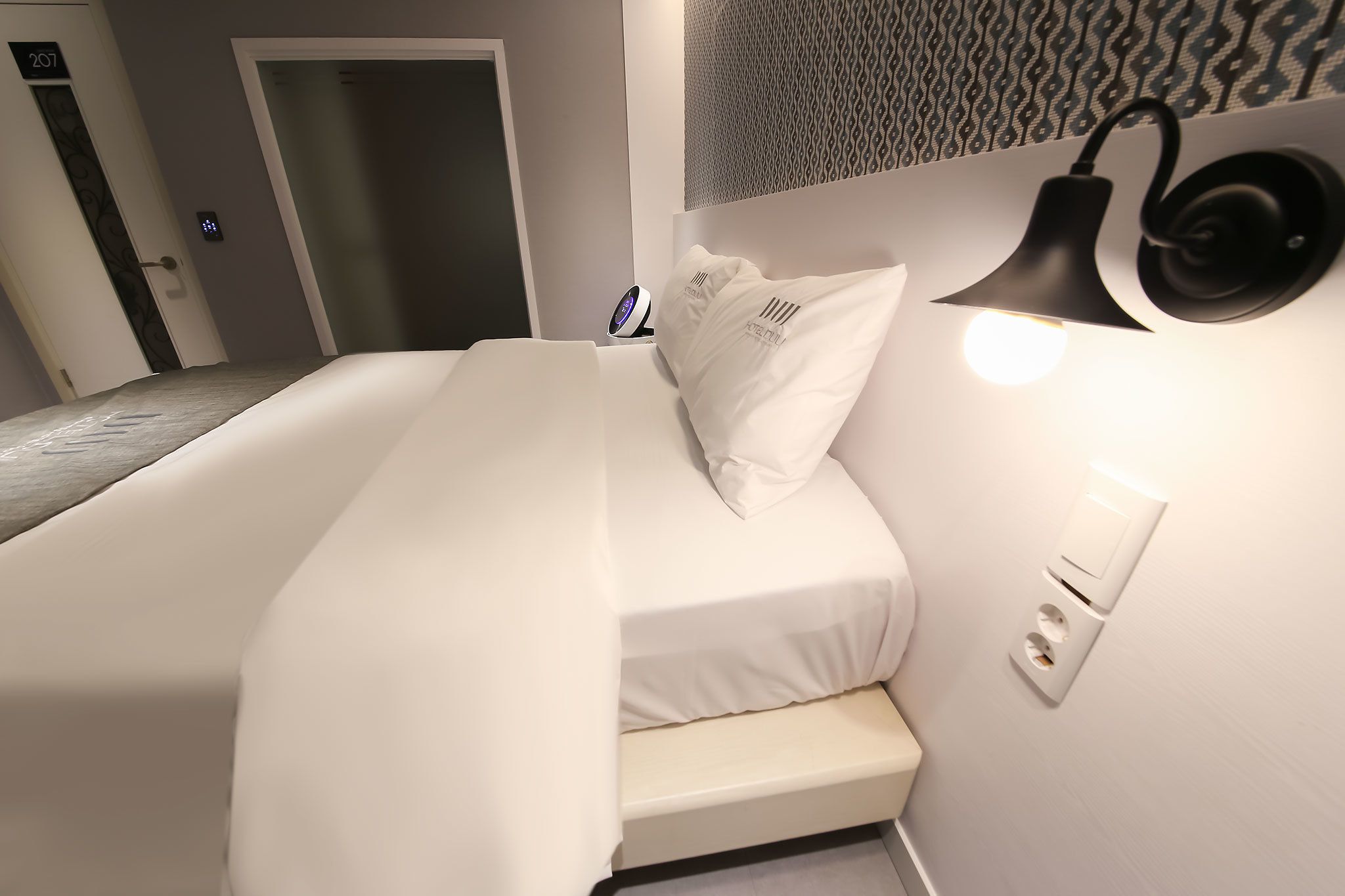 Una habitación de hotel con cama, mesita de noche y lámpara de pared.