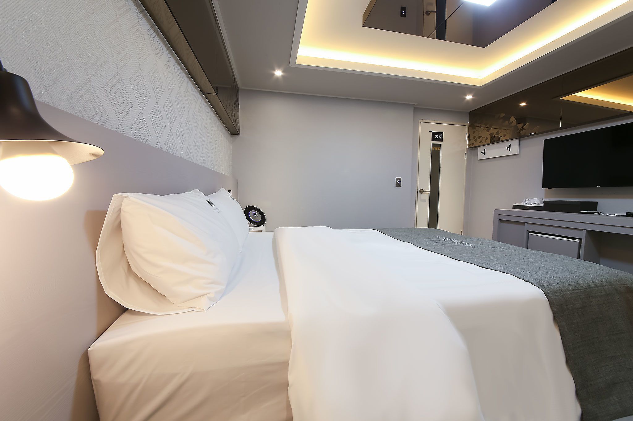 A modern hotel room with a neatly made bed, a bedside table with a lamp, a flat-screen TV, and a desk with a chair.