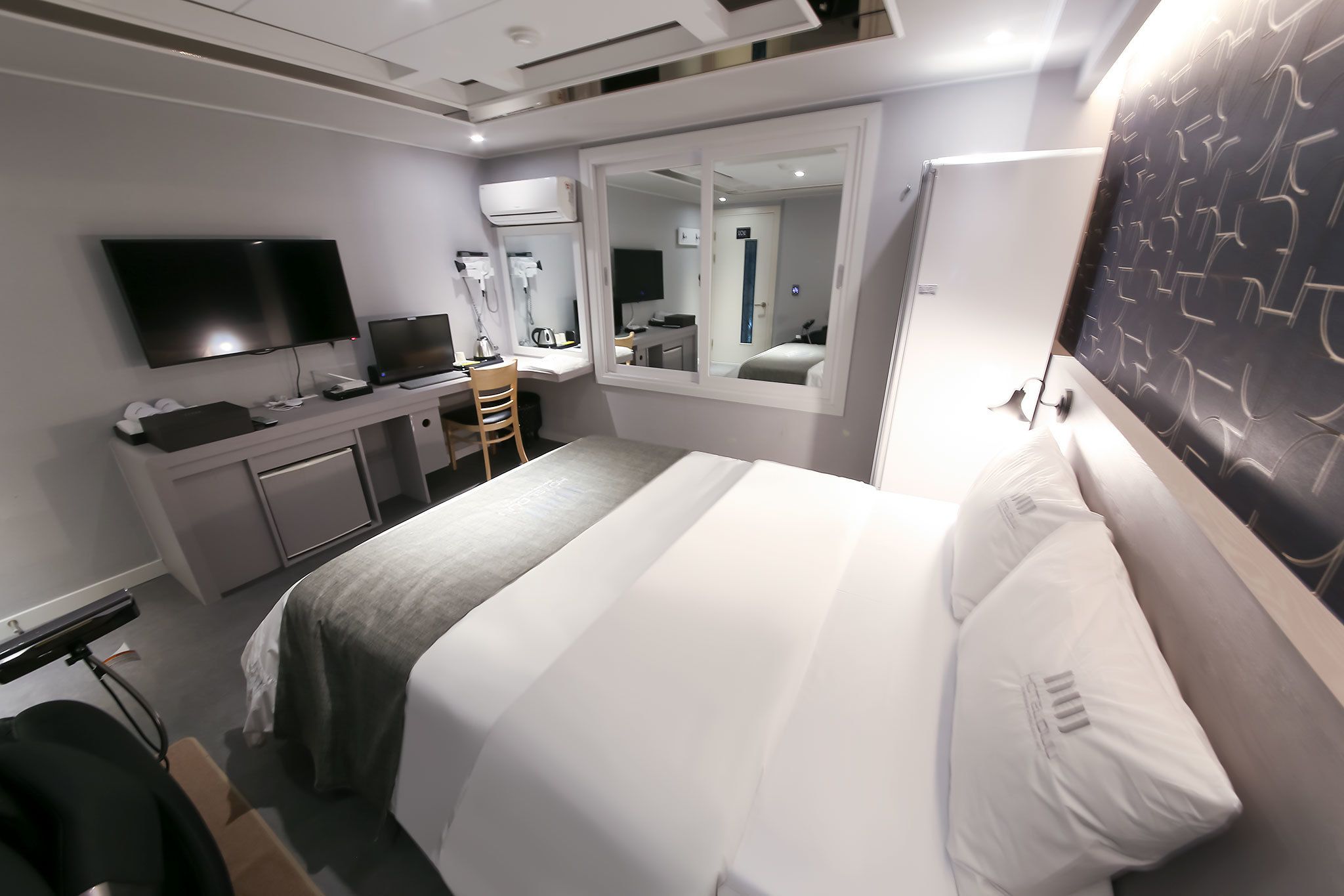 Habitación de hotel con cama, escritorio, computadora, televisor y espejo.
