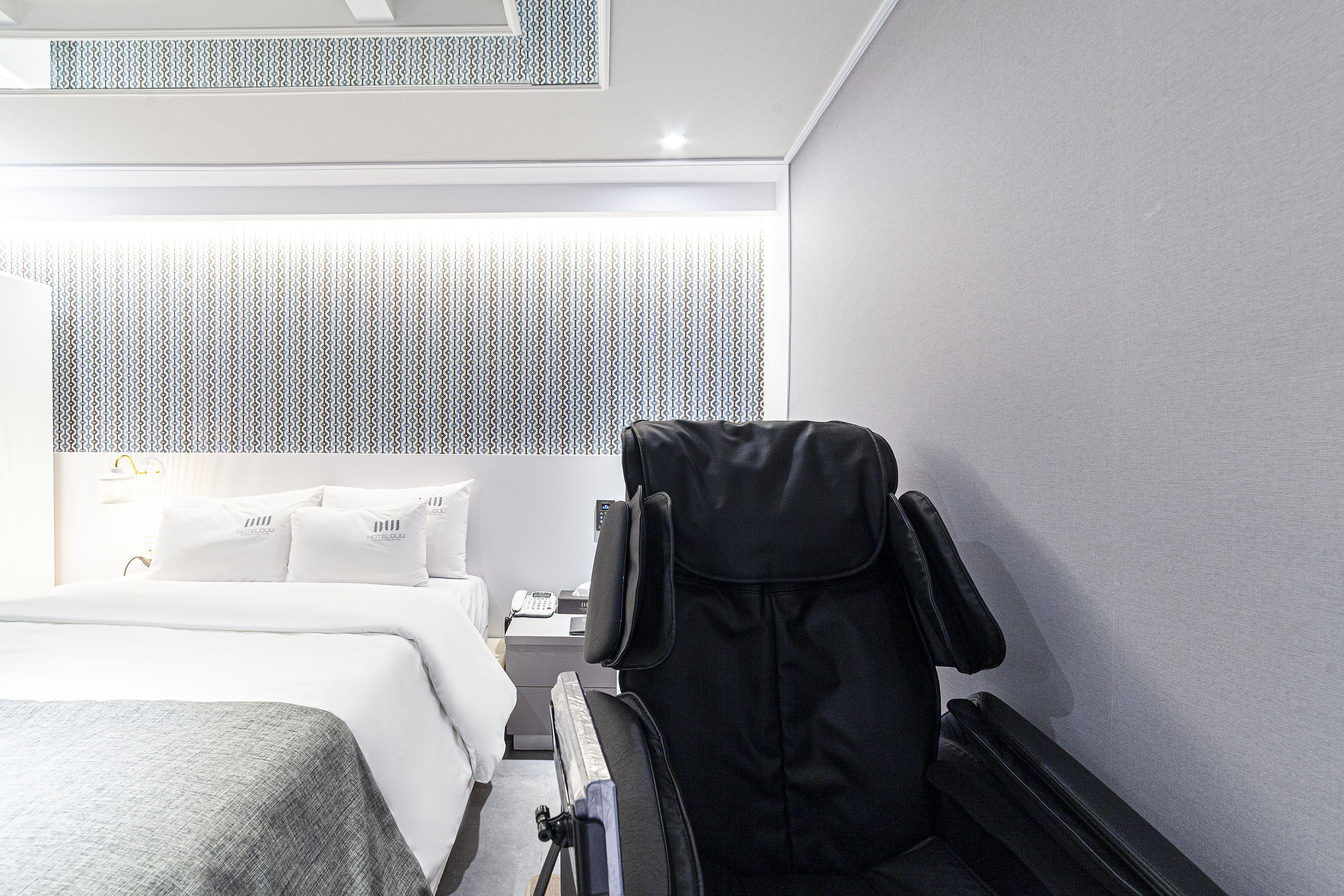 Una habitación de hotel moderna con una cama bien tendida, un sillón de masaje y una mesita de noche con lámpara.