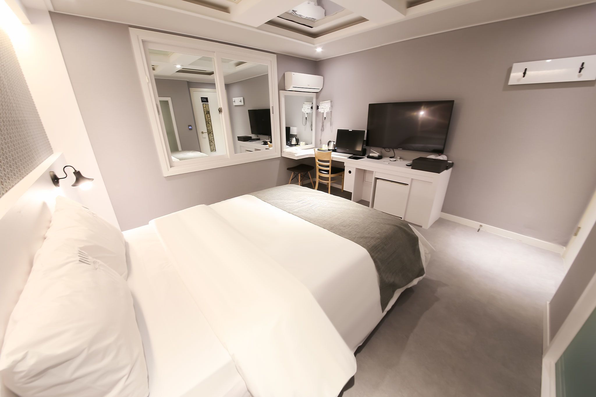 A modern hotel room with a large bed, a desk with a computer, a TV, and a mirror.