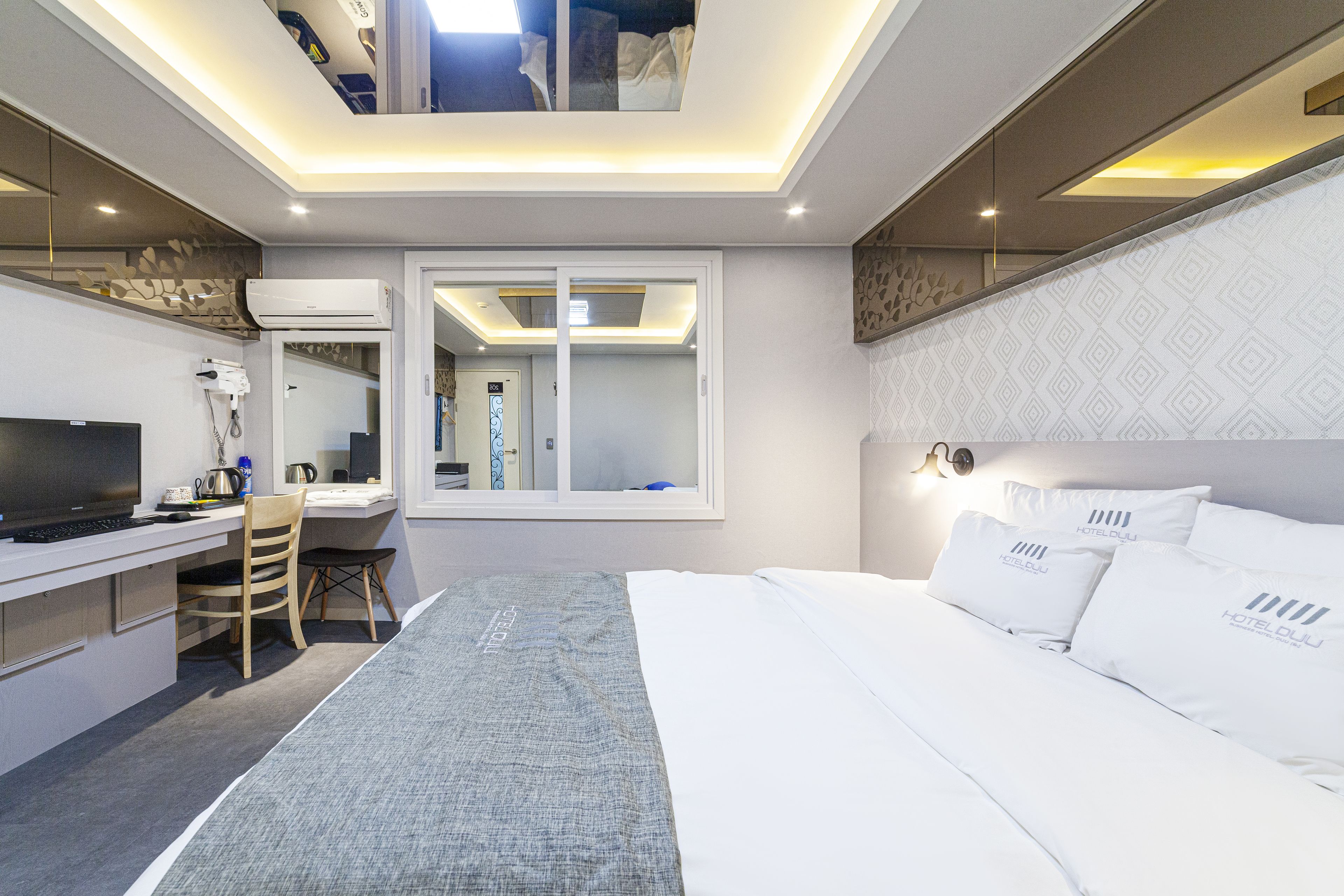 A modern hotel room with a bed, desk, and chair, a large window, and a ceiling with recessed lighting.