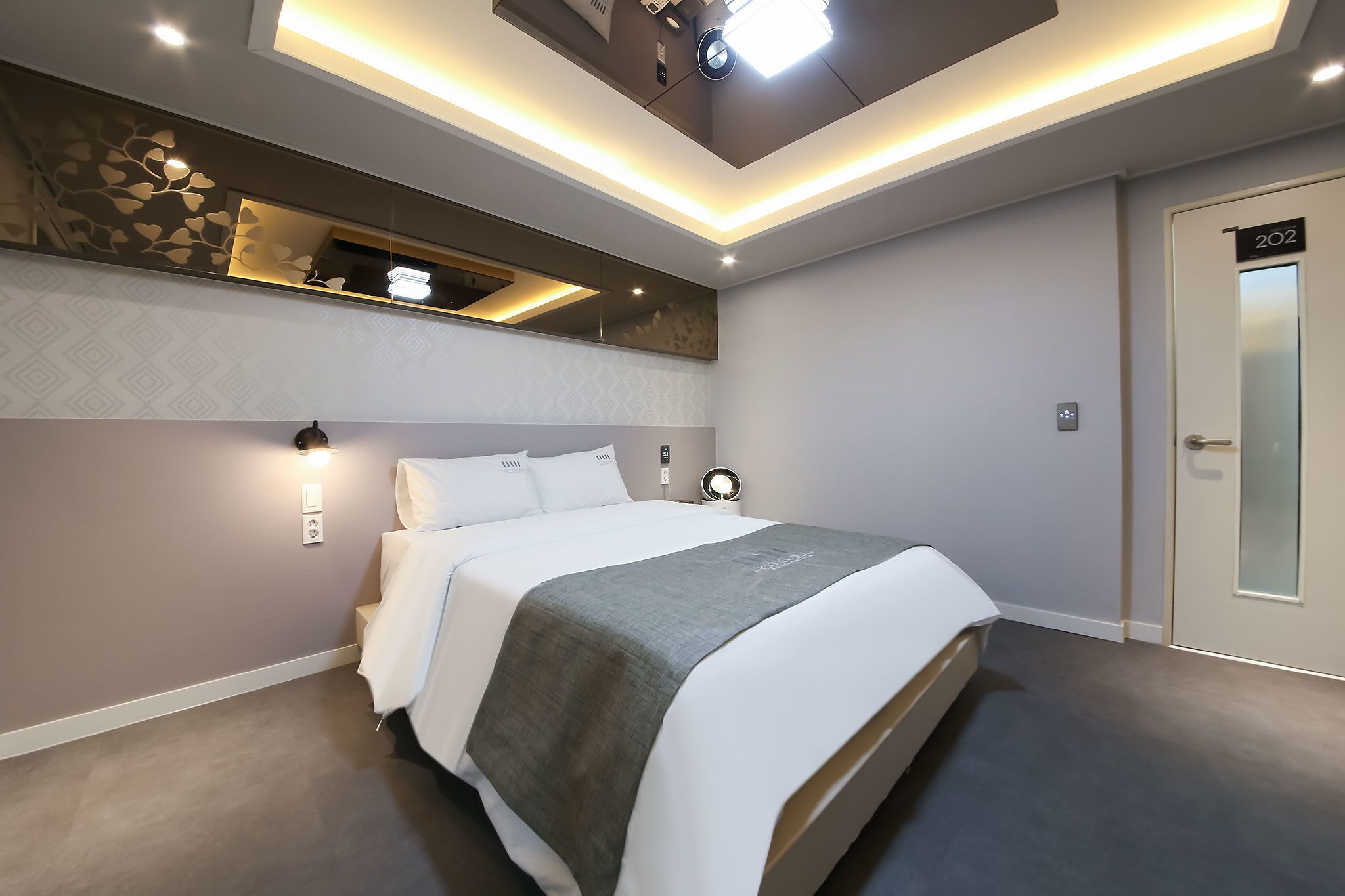 A modern hotel room with a neatly made bed, a small seating area with a chair and table, a built-in shelf with decorative items, and a door marked with the number 202.