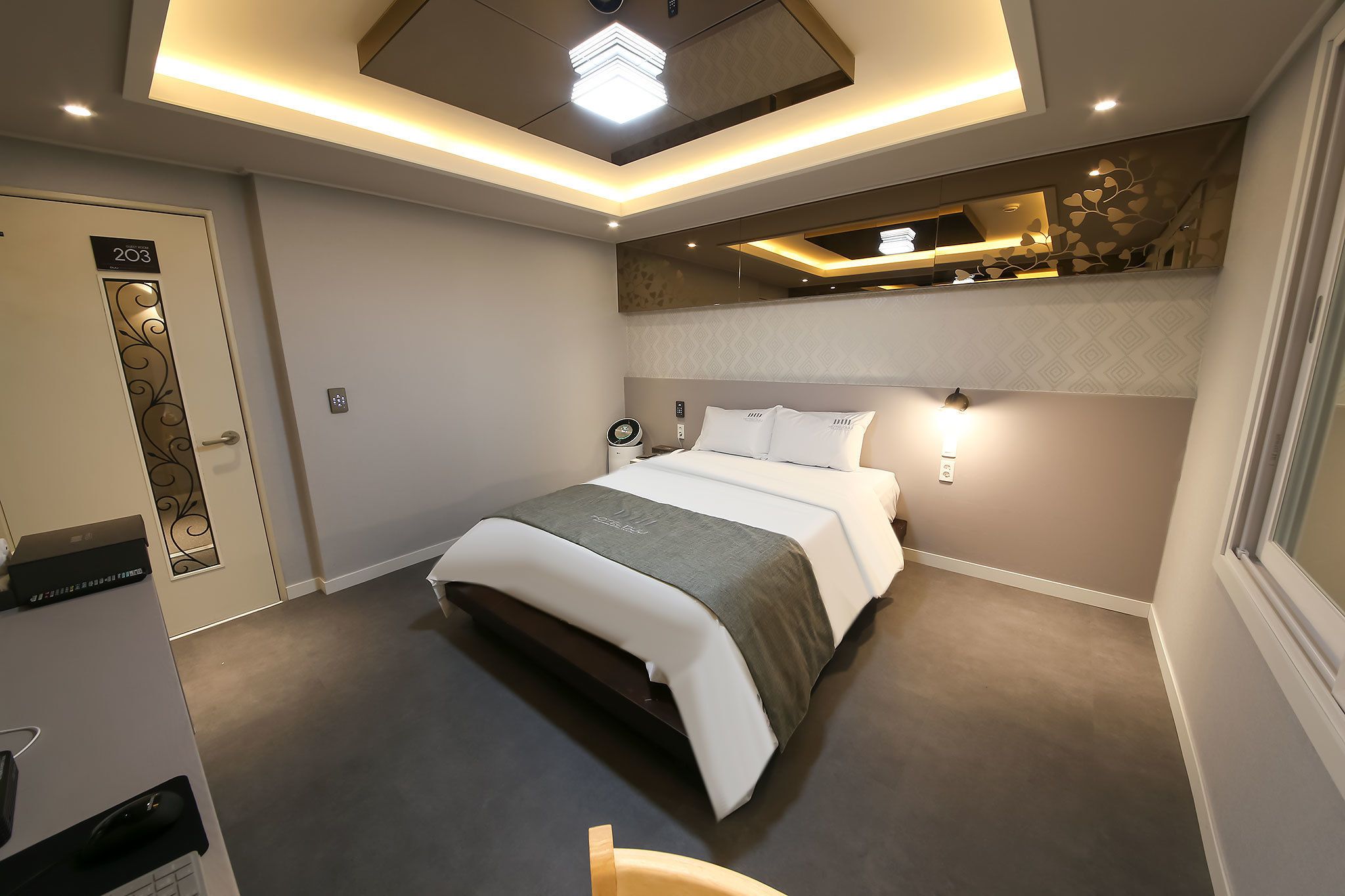 A modern hotel room with a neatly made bed, a desk with a computer, and a ceiling light fixture.