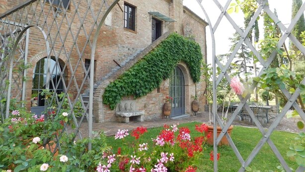 Un bâtiment en briques avec un banc en pierre, une treille en métal et une variété de fleurs au premier plan.