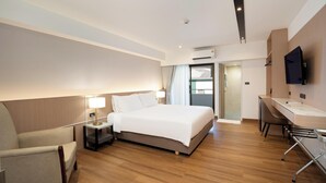 Quarto de hotel moderno com cama, mesa de cabeceira, luminária, poltrona, escrivaninha e televisão.
