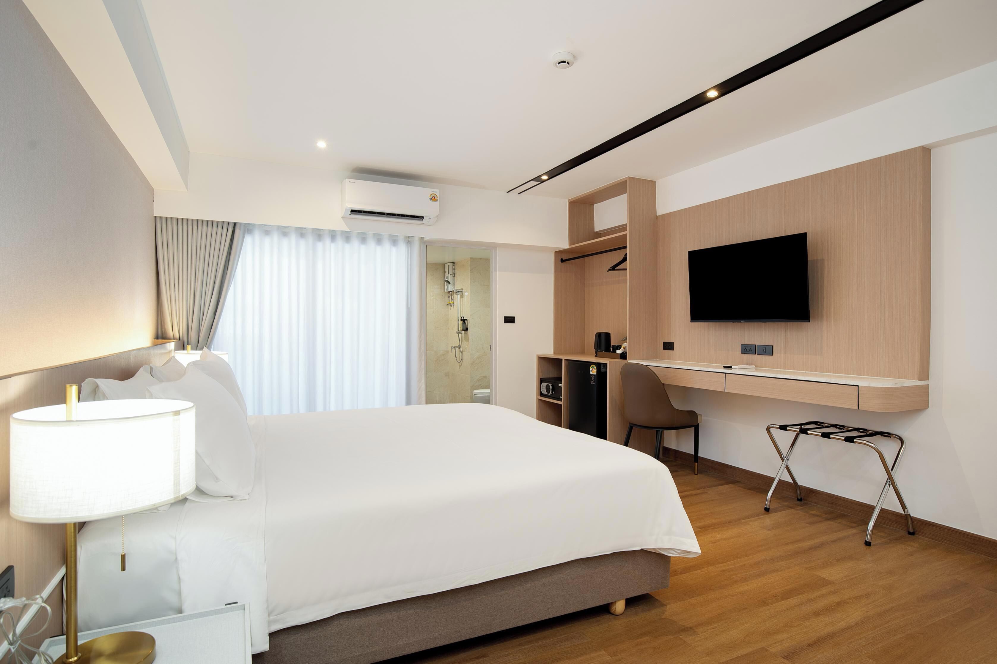 A modern hotel room with a bed, bedside lamp, desk, chair, TV, and built-in wardrobe.
