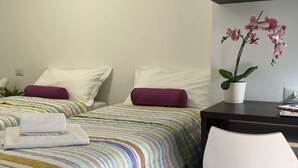 A hotel room with two beds, each with a striped bedspread and pillows, a small desk with a potted orchid, and a white chair.