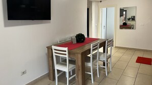 A dining area with a wooden table, white chairs, a red placemat, and a television mounted on the wall.