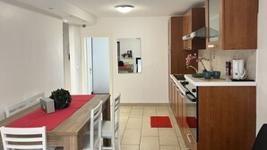 A compact kitchen with wooden cabinets, a dining table with chairs, and a small countertop with a kettle and cups.