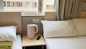 A hotel room with two beds, a nightstand with a kettle and remote control, and a window with curtains.