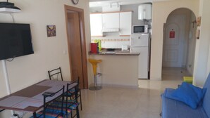 Een eetgedeelte met een tafel en stoelen, een keuken met kasten en apparatuur, en een woonkamer met een bank.
