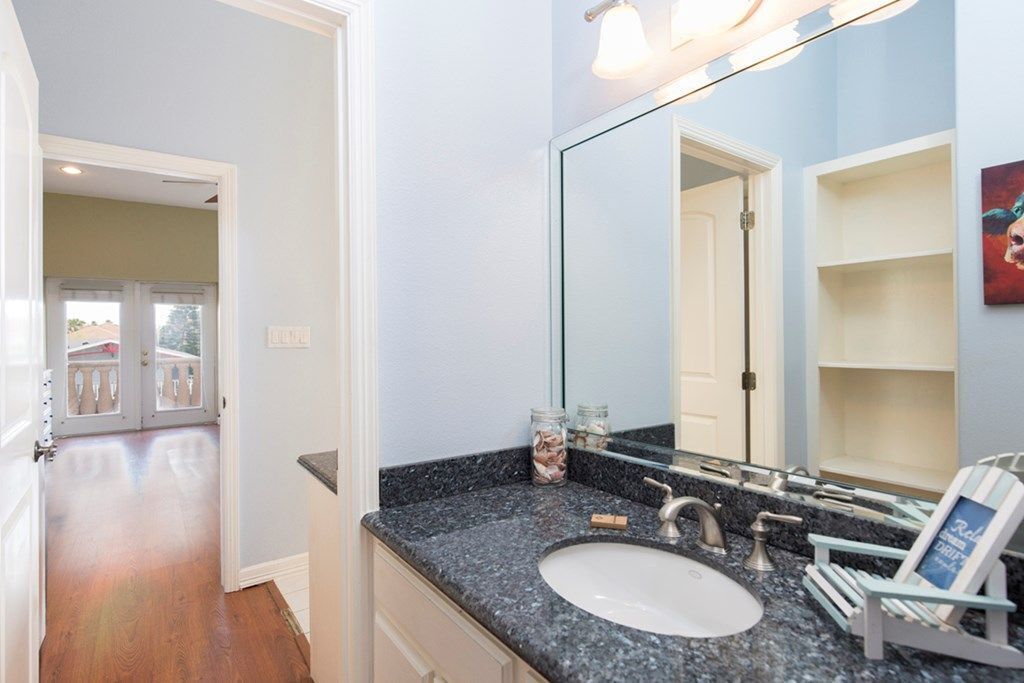 Ein Badezimmer mit einer Granit-Arbeitsplatte, einem weißen Waschbecken und einem großen Spiegel. Eine Tür führt in einen anderen Raum, und ein Regal trägt einen Bilderrahmen.