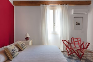 Un dormitorio con cama, mesita de noche, lámpara, ventana con cortinas y una silla roja.