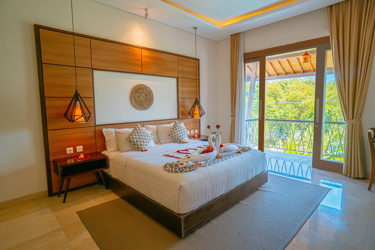 A spacious bedroom with a large bed, a wooden headboard, and a balcony with a view of greenery.