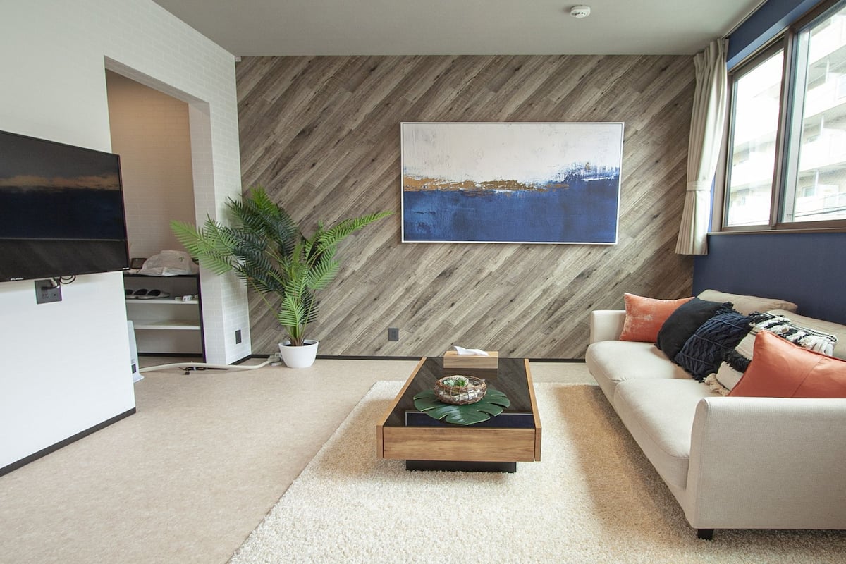 Ein modernes Wohnzimmer mit Flachbildfernseher, einer Wandpaneele aus Holz, einer Couch mit Kissen, einem Couchtisch und einer Topfpflanze.