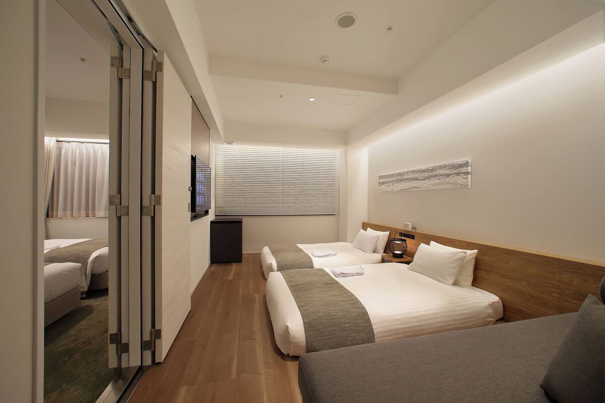 A hotel room with two beds, a wooden headboard, a large window with blinds, a flat-screen TV, and a small bedside table.