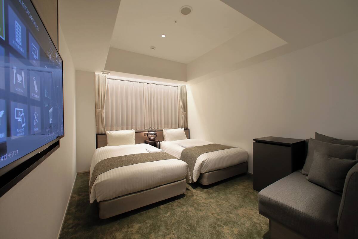 A hotel room with two beds, a desk, a chair, and a large screen displaying room information.