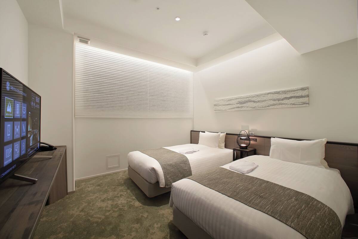 A hotel room with two beds, a TV, a window with blinds, and a wall-mounted artwork.